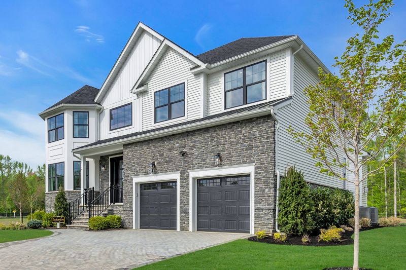 Exterior photo of the Lockwood model at the Grove at Woodhaven showing another angle of a modern two-story suburban house with a combination of white siding and gray stone exterior. Large, black-trimmed windows are symmetrically placed across. The front features a large, black-framed glass door, two black garage doors with rectangular paneling, and a prominent bay window section on the left side of the house. The home is surrounded by neatly landscaped greenery, including small shrubs, young trees, and a well-maintained lawn.