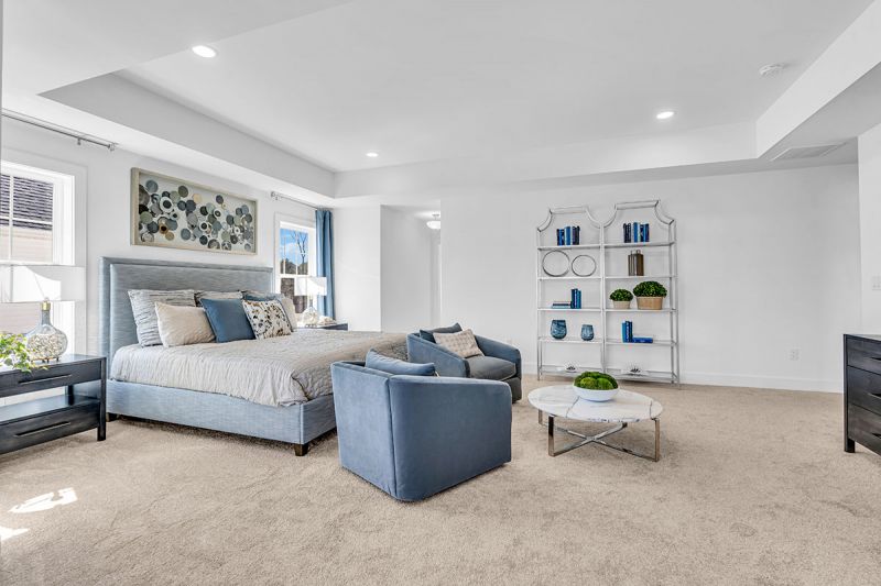 Interior photo of the Boulder model at the Grove at Woodhaven showcasing the master bedroom featuring wall-to-wall carpeting, a bed with two nightstands on each side, dresser and two accent chairs with table in front. There are large windows to allow for natural light.