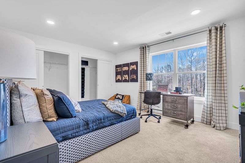 Interior photo of the Boulder model at the Grove at Woodhaven showcasing the fourth bedroom with wall-to-wall carpeting, a bed with two nightstands, a dresser, and a study table with a rolling chair. There is a double closet with ample storage . A large window brings in plenty of natural light.