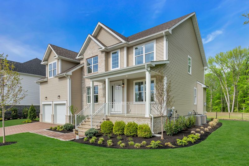 Exterior photo of the Boulder model at the Grove at Woodhaven showing another angle of a modern two-story house with beige siding and light brick accents. The home features a covered front porch with white railings, a white front door at the top of brick steps, and a symmetrical arrangement of white-trimmed windows. There are two white garage doors on the left side, and the roof has brown shingles with gabled dormers. The home is surrounded by neatly landscaped greenery, including small shrubs, young trees, and a well-maintained lawn.