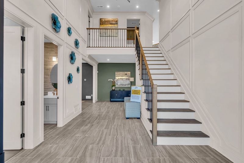 Interior photo of the Lockwood model at the Grove at Woodhaven, showcasing the front entrance featuring luxury grey oak flooring, modern staircase with railings leading to the top floor and squared paneled walls with high vaulted ceilings. A spacious hallway connects to other areas of the house.