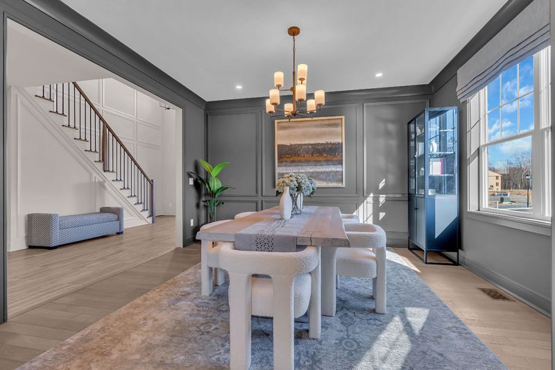 Interior photo of the Lockwood model at the Grove at Woodhaven, showcasing the dining room with luxurious oak flooring. An elegant chandelier hangs above a six-person table surrounded by chairs. The space features elegant decor and artwork. A staircase leading to the upper floor is visible in the background.