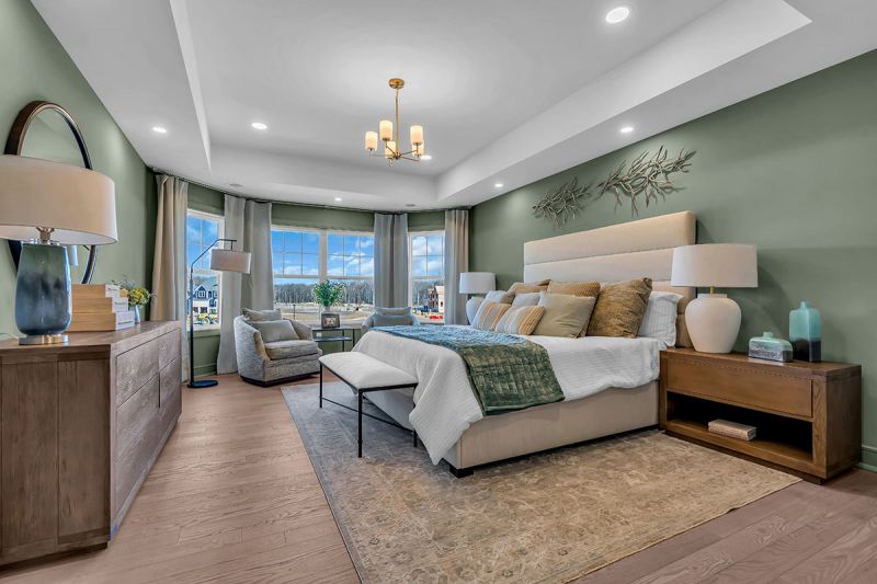 Interior photo of the Lockwood model at the Grove at Woodhaven showing the master bedroom featuring luxury oak flooring, a bed with two nightstands on each side, a dresser and accent chair. There is a large window for natural light.