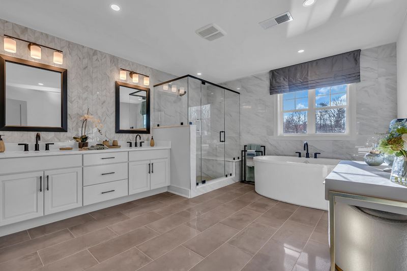 Interior photo of the Lockwood model at The Grove at Woodhaven showcasing the master bathroom with luxury slate tile flooring, a double-sink vanity with a mirror and multiple light fixtures above, a stand-up shower, and a freestanding tub. A large window fills the space with natural light.