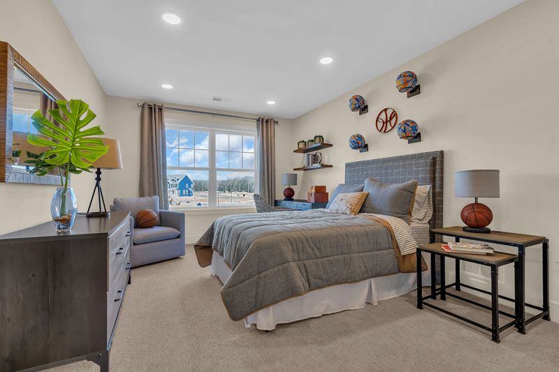 Interior photo of the Lockwood model at the Grove at Woodhaven showing the third bedroom with wall-to-wall carpeting, a bed with a nightstand, a dresser, and a study table with a rolling chair. A large window fills the room with natural light.