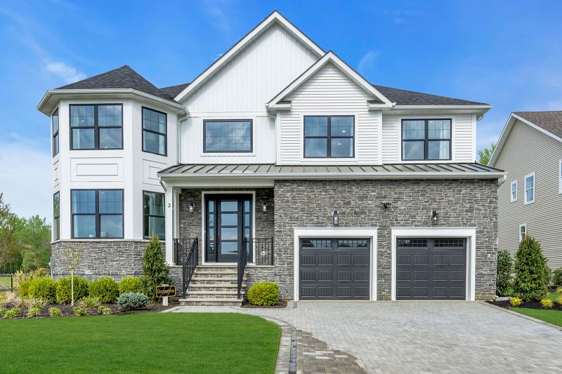 Exterior photo of the Lockwood model at the Grove at Woodhaven showing a modern two-story suburban house with a combination of white siding and gray stone exterior. Large, black-trimmed windows are symmetrically placed across. The front features a large, black-framed glass door, two black garage doors with rectangular paneling, and a prominent bay window section on the left side of the house. The home is surrounded by neatly landscaped greenery, including small shrubs, young trees, and a well-maintained lawn.