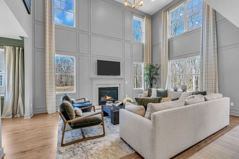 Interior photo of the Sequoia model at the Grove at Woodhaven showing another angle of the family room featuring luxury wood flooring, high vaulted ceilings with large windows throughout the unit. A large L- shaped couch with an ottoman and two accent chairs can be seen in the center of the room. A fireplace and mounted T.V complete the space.