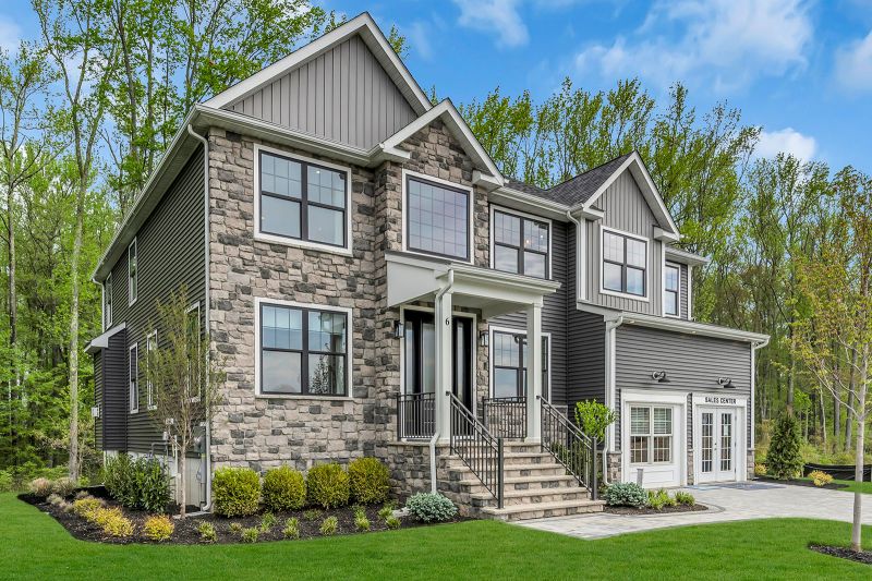 Exterior photo of the Sequoia model at the Grove at Woodhaven showing a modern house with a combination of stone and gray siding exterior. Large, black-trimmed windows are symmetrically placed across. The front entrance includes stone steps, white columns and black metal railing. To the right is a two-car garage with stone steps that lead to the main road. The home is surrounded by neatly landscaped greenery, including small shrubs, young trees, and a well-maintained lawn.