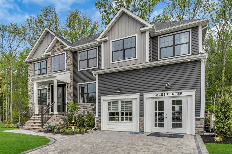 Exterior photo of the Sequoia model at the Grove at Woodhaven showing the backside of a modern house with a combination of stone and gray siding exterior. Large, black-trimmed windows are symmetrically placed across. A raised back deck with white railings and glass sliding doors extends from the house.  The home is surrounded by neatly landscaped greenery, including small shrubs, young trees, and a well-maintained lawn.